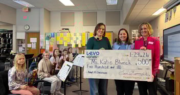 Three women hold a $500 check for Adopt A Classroom with students in the background at a Sioux Falls school