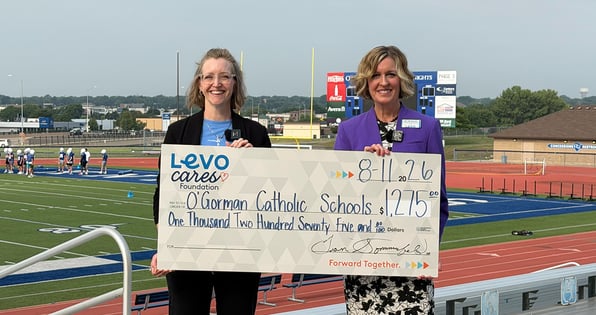 two women hold a check for O'Gorman Catholic Schools Foundation in front of a football field